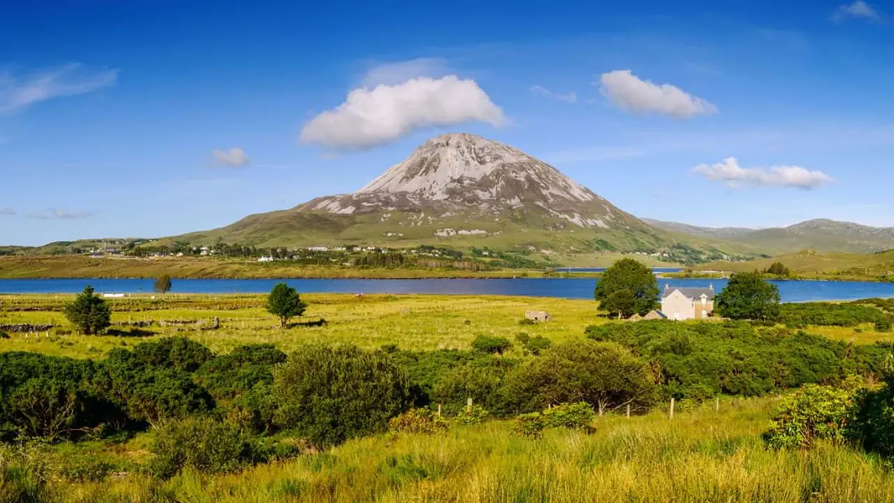 View of a mountain with fields in the forefront, and a river running in between. Blue sky with some clouds