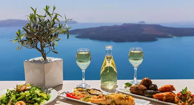 Three plates of Greek food, and 2 glasses and a bottle of wine, with view out to Greek Islands