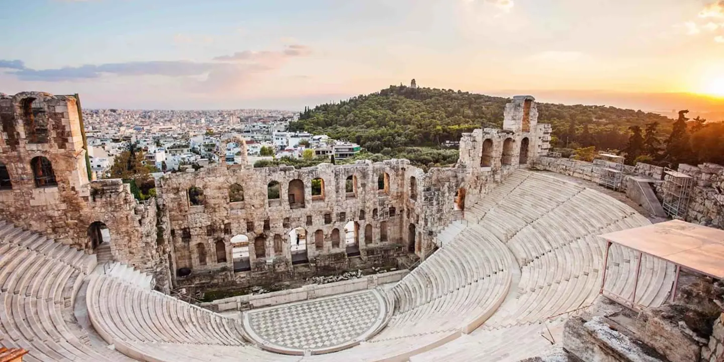 High angle view of the ruins of an amphitheatre, with the view of a city in the distance and forested land to the left.