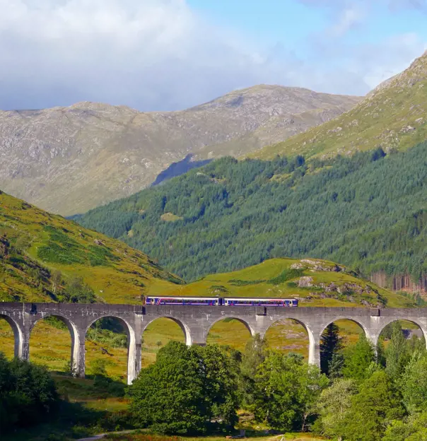 A train going along Glenfinnan Viaduct Bridge with the view of the mountains behind 