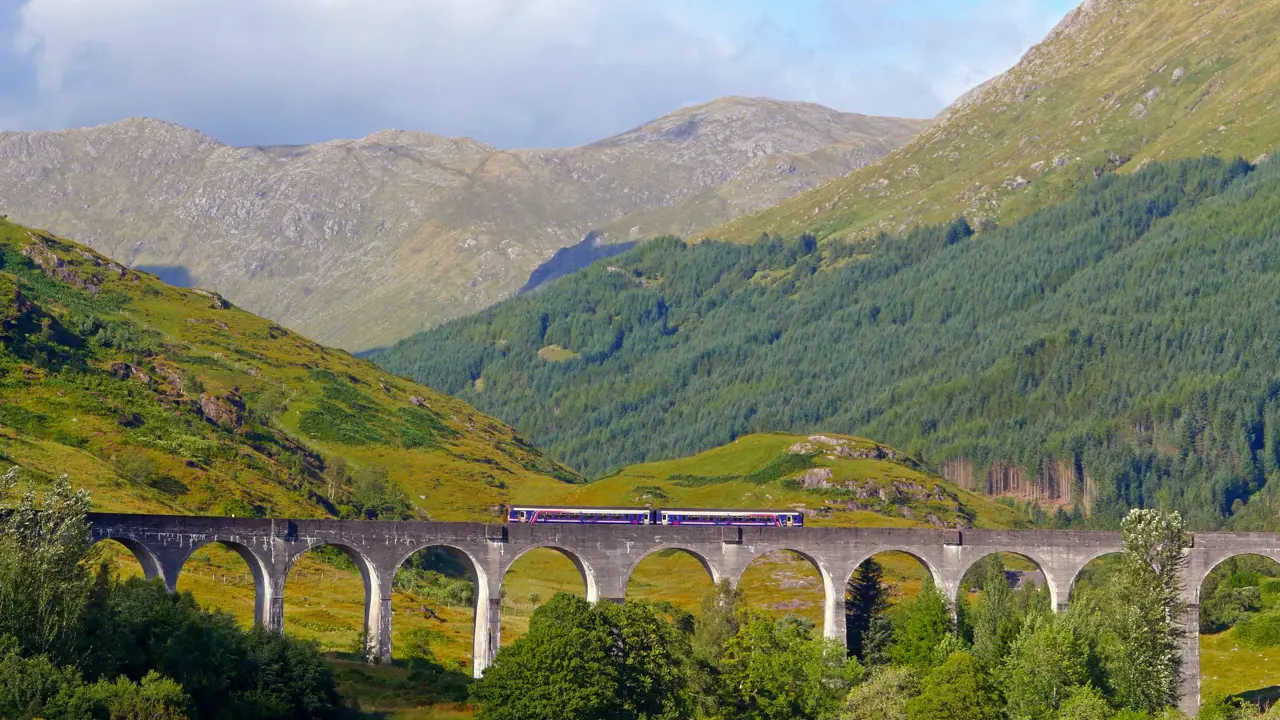 A train going along Glenfinnan Viaduct Bridge with the view of the mountains behind 