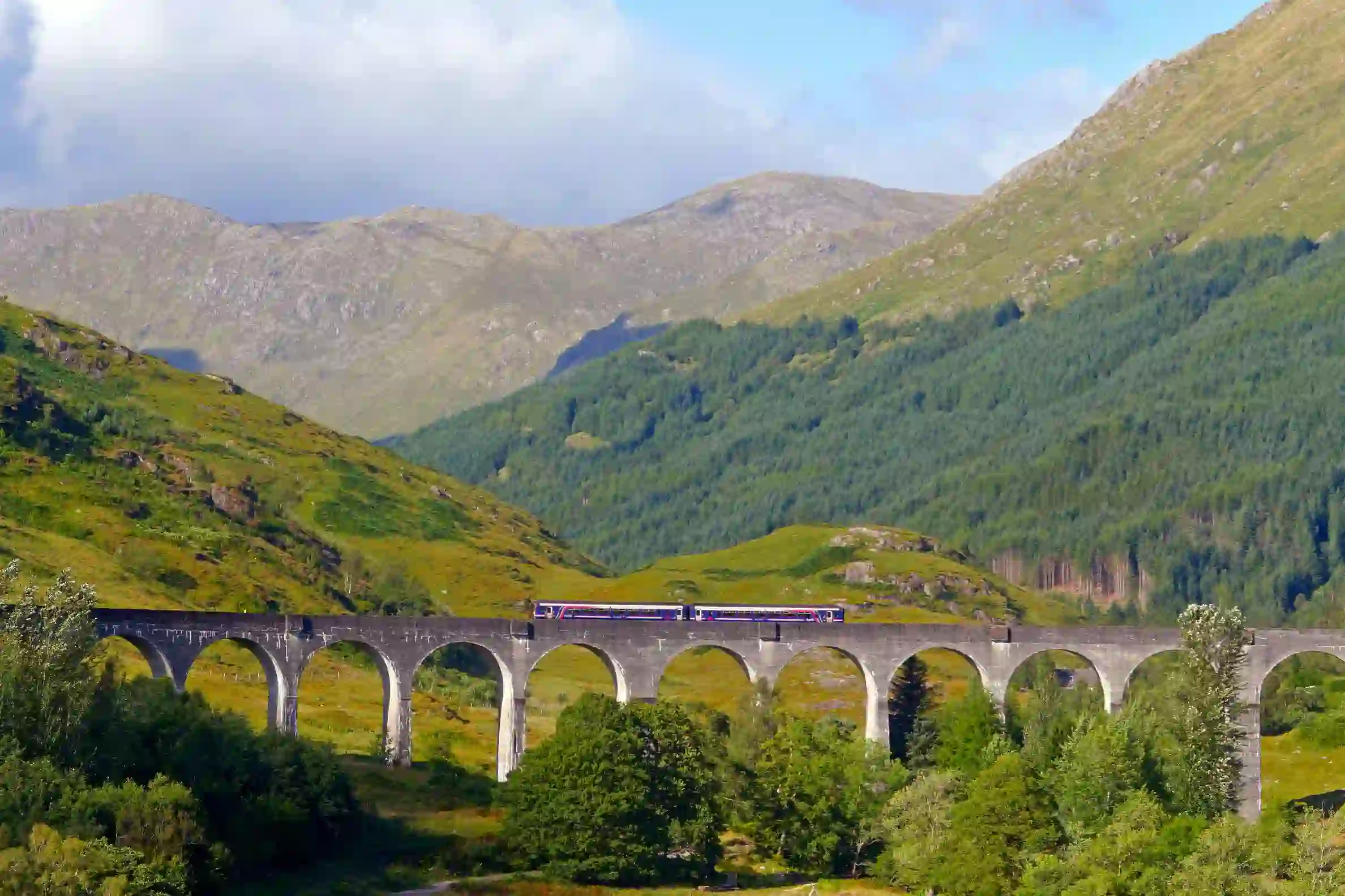 A train going along Glenfinnan Viaduct Bridge with the view of the mountains behind 