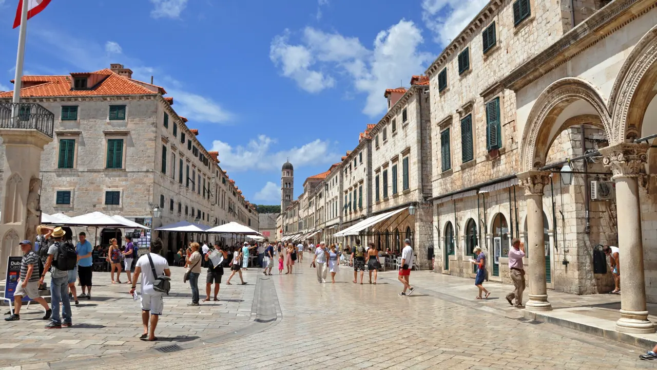 View of a street in the Old Town, Dubrovnik, bricked buildings with orange roofs either side and people in the street. White umbrellas all down the road an a tower at the end. Blue sky with some clouds.