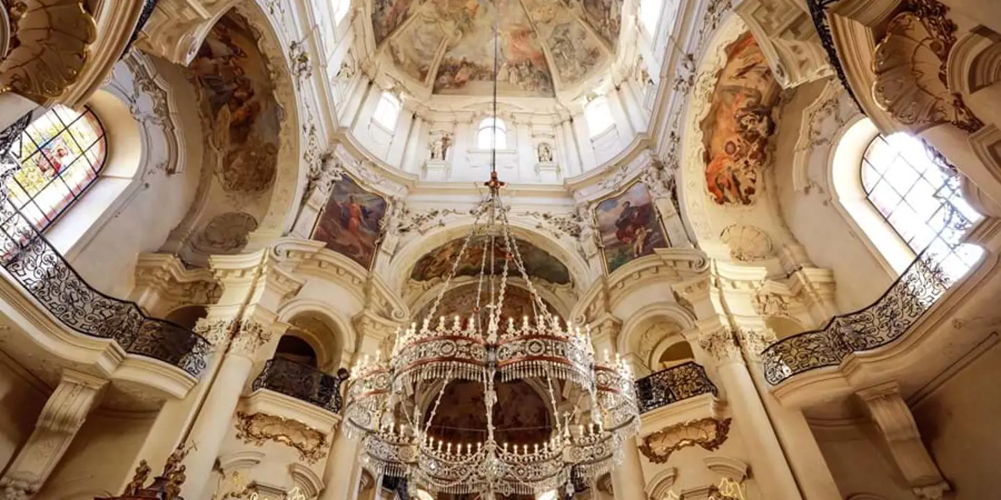 Under shot of a ceiling and chandelier, with old religious artwork and architectural design.