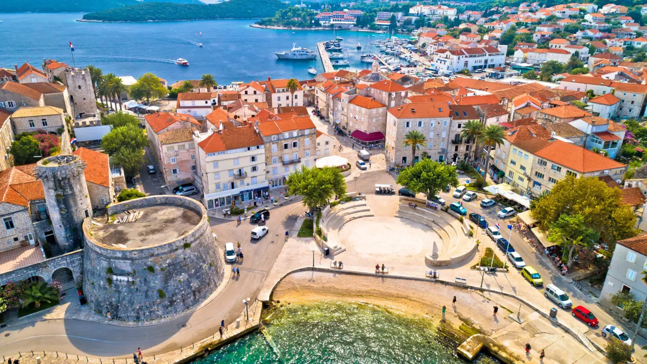 Aerial view of a town, with cream buildings with orange roofs, going into the distance on the right with, overlooking the sea on the left. In the forefront a circular stone building, and a circular seating area, surrounded by cars and some pedestrians. Slight view of water also in the forefront. 