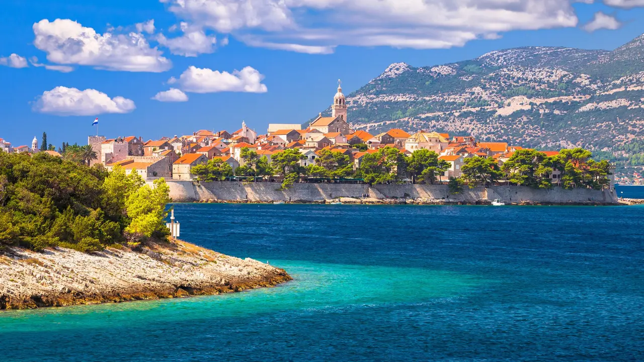 View of an island from the sea. In the forefront to the left, there is a piece of land with bushes, and in the centre, houses with orange roofs and a tower in the middle. To the right of the image, a mountain with some forest and spaces of land on, and a blue sky.