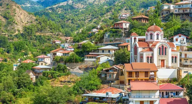 View of a town in the hills amongst the trees, with houses with orange and red roofs and a orange and white church to the right with curved roofs and a dome on top with a cross on. To the left is the hills behind, covered in trees.