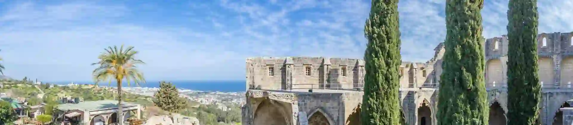 Stone ruins to the right, with a grassy area in the middle with three tall slim trees. Opposite, to the left is more grassy area with a palm tree and bushes. Behind this is a distant view of a town and the ocean, and a blue sky above.