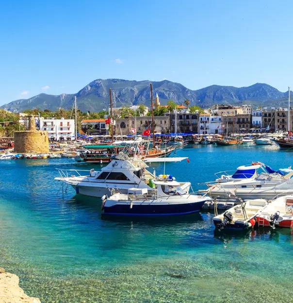 Boats docked at a harbour with bright blue water, buildings on the other side, and ruins of a castle to the left. Mountains in the distance in front of a blue sky.