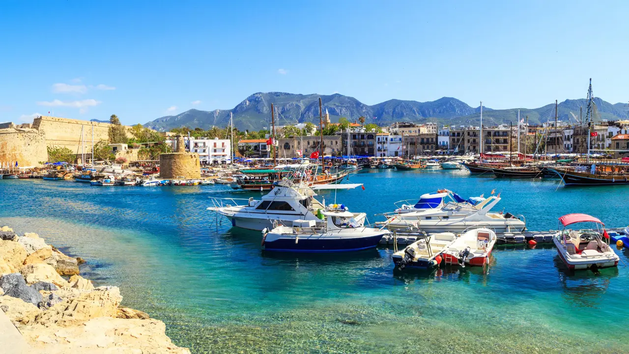 Boats docked at a harbour with bright blue water, buildings on the other side, and ruins of a castle to the left. Mountains in the distance in front of a blue sky.