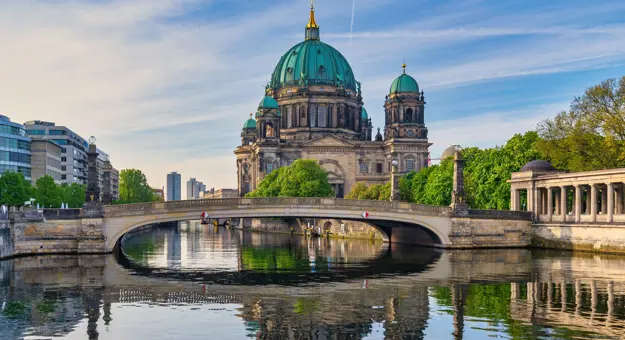 View of Berlin Cathedral, which is grey with turquoise domed roofs, and has a gold turret in the middle. In front is a bridge, and there is water in the forefront