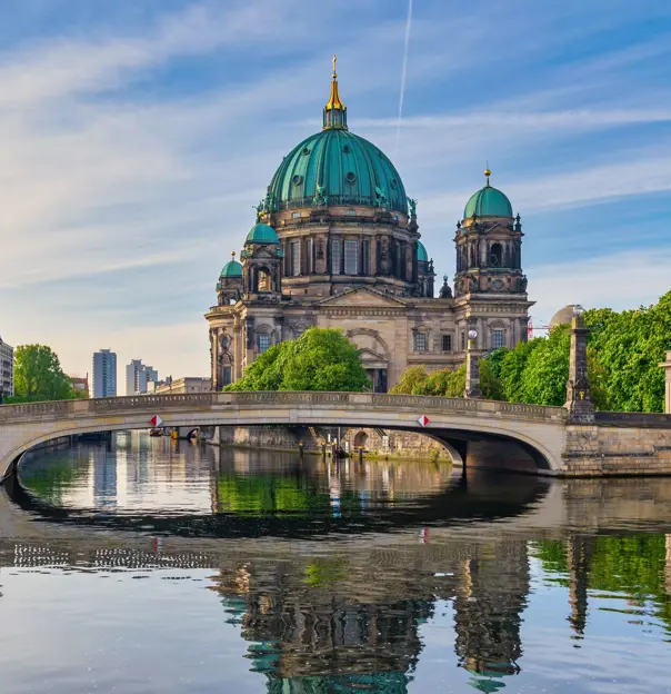 View of Berlin Cathedral, which is grey with turquoise domed roofs, and has a gold turret in the middle. In front is a bridge, and there is water in the forefront