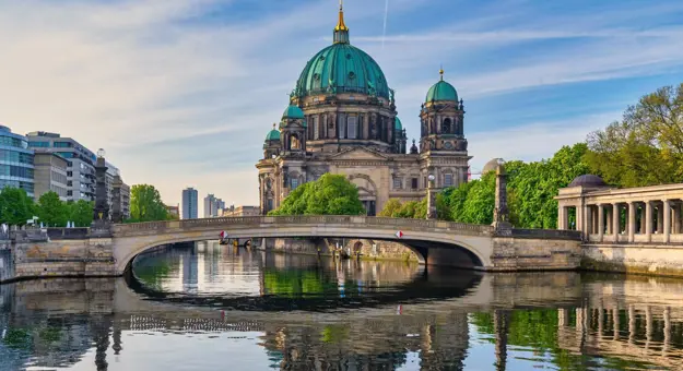 View of the Berlin Cathedral (Berliner Dom) with its green dome reflected in the River Spree, framed by a stone bridge, historic colonnades, and modern city buildings under a lightly clouded sky