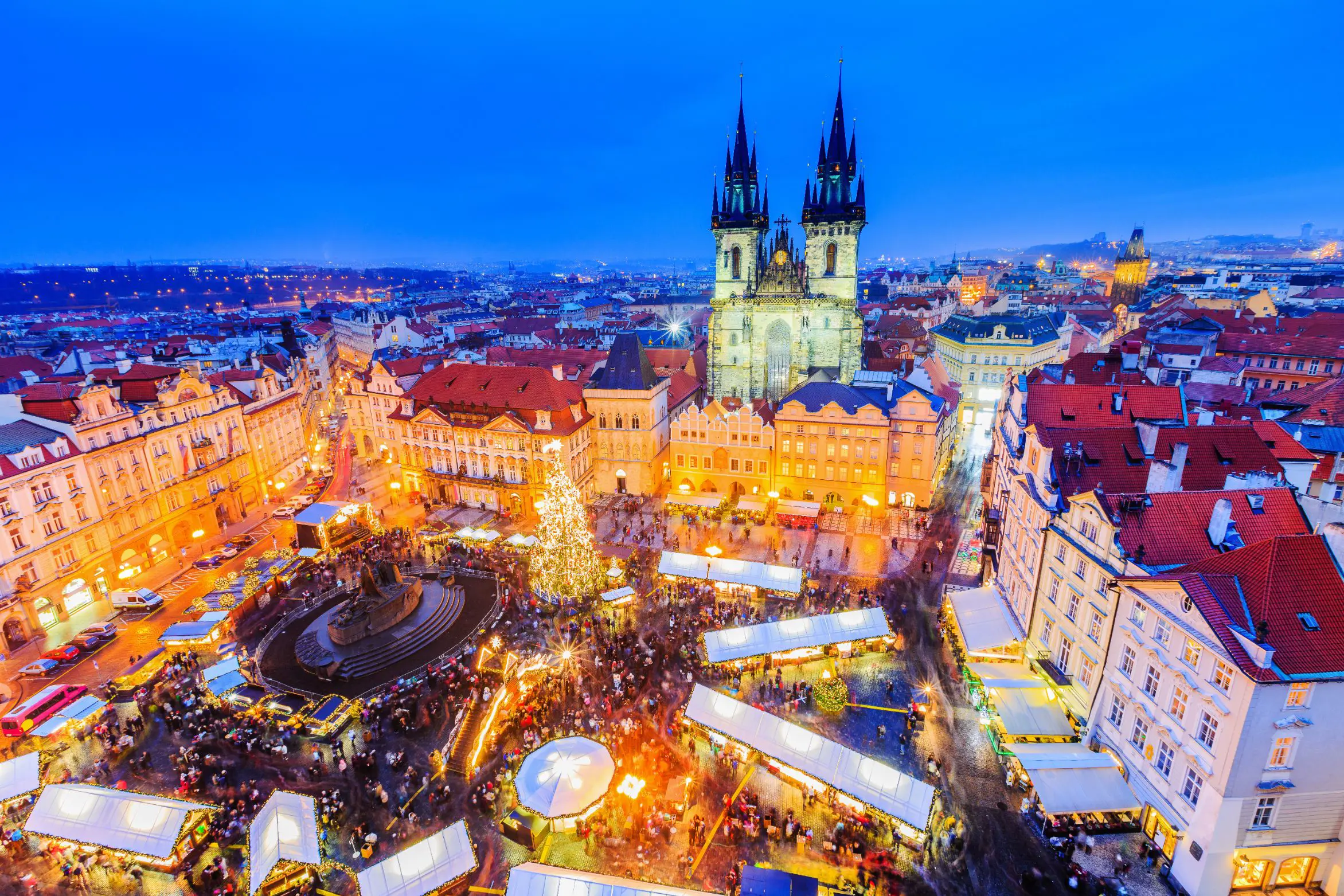 High angle shot of Christmas market in the town square of Prague, showing a lit up gothic church with two towers and spiky turrets, a monument with a circular barrier around it in the centre of the square, next to a large lit up Christmas tree. White roofs of the stalls are all around the square surrounded by people. 