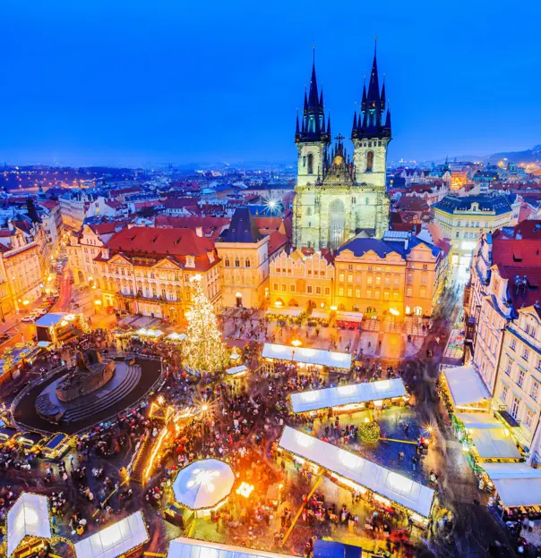 High angle shot of Christmas market in the town square of Prague, showing a lit up gothic church with two towers and spiky turrets, a monument with a circular barrier around it in the centre of the square, next to a large lit up Christmas tree. White roofs of the stalls are all around the square surrounded by people. 