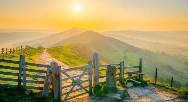 View of the Peak District at sunrise, with a gate at the forefront of the image and a trail that leads all the way to the mountains in the far distance