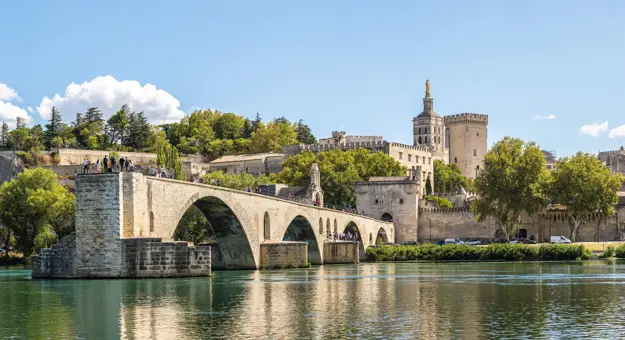 Wide angle shot of the Pont d'Avignon bridge, with the palace on the other end. The river water in the forefront, and a blue sky with clouds above.