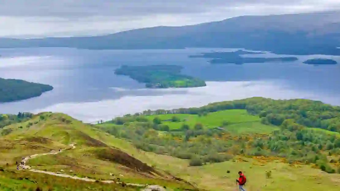 High angle view of Loch Lomond and Trossachs National Park, with a man wearing a red jacket walking down the mountain