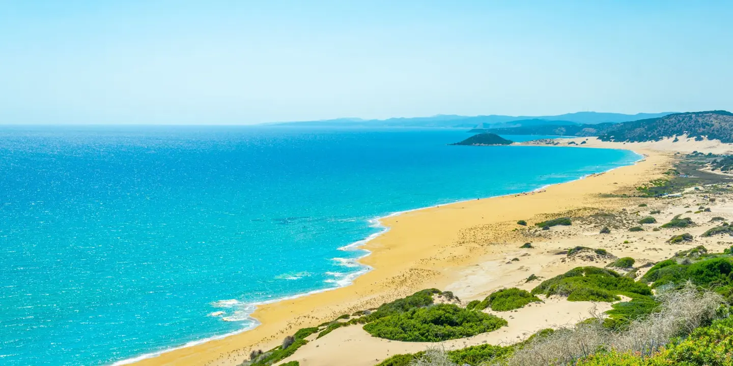 High angle shot of a strip of beach, with yellow sand and bright blue water, leading towards mountains in the distance. The land behind the beach the right forefront has patches of grass and sand.