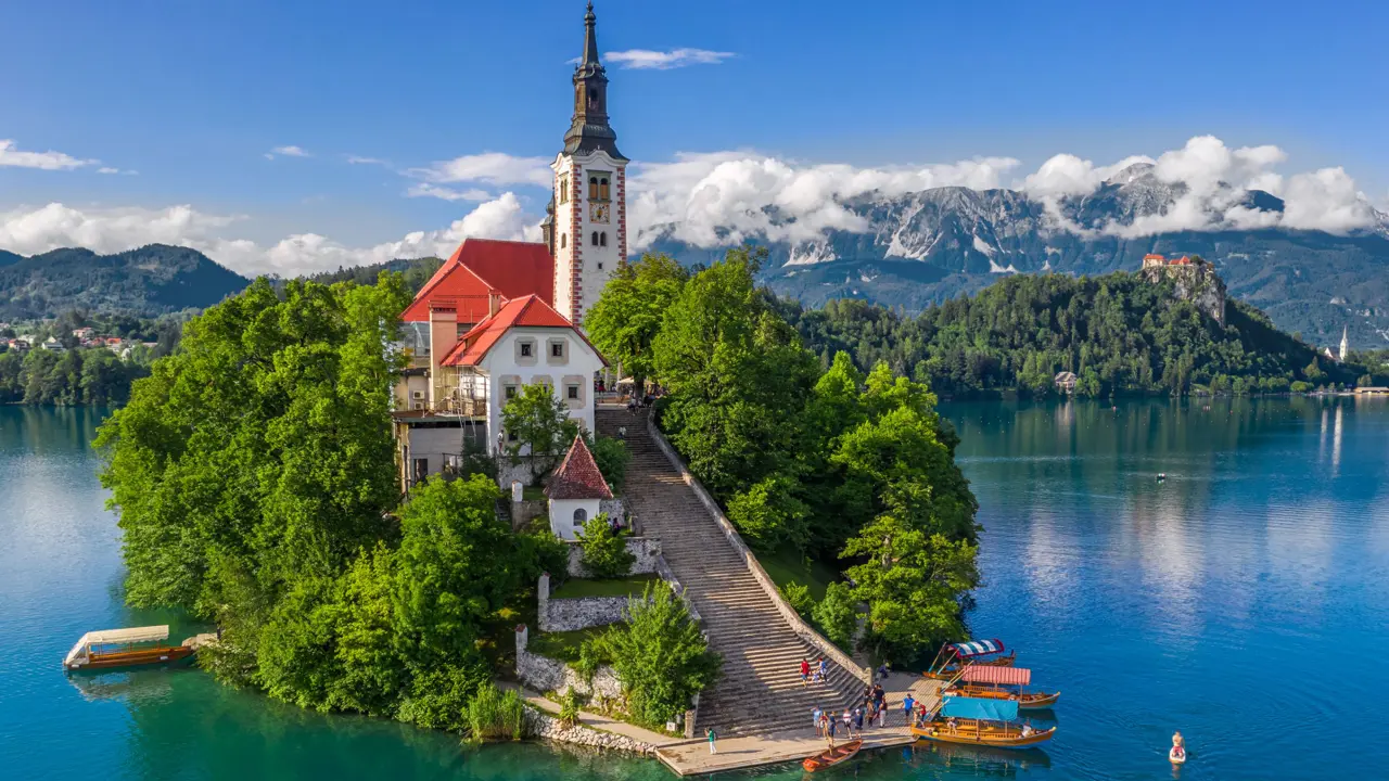 Shot of a church on a small bit of forested land on water, there is a pathway leading up to the church which has some people and boats at the bottom. Behind, there are mountains and clouds, under a blue sky. 