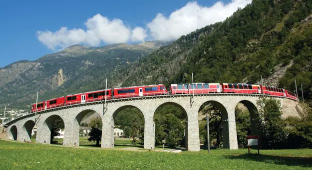 The Bernina Express going across a bridge, with grassy mountains in the background