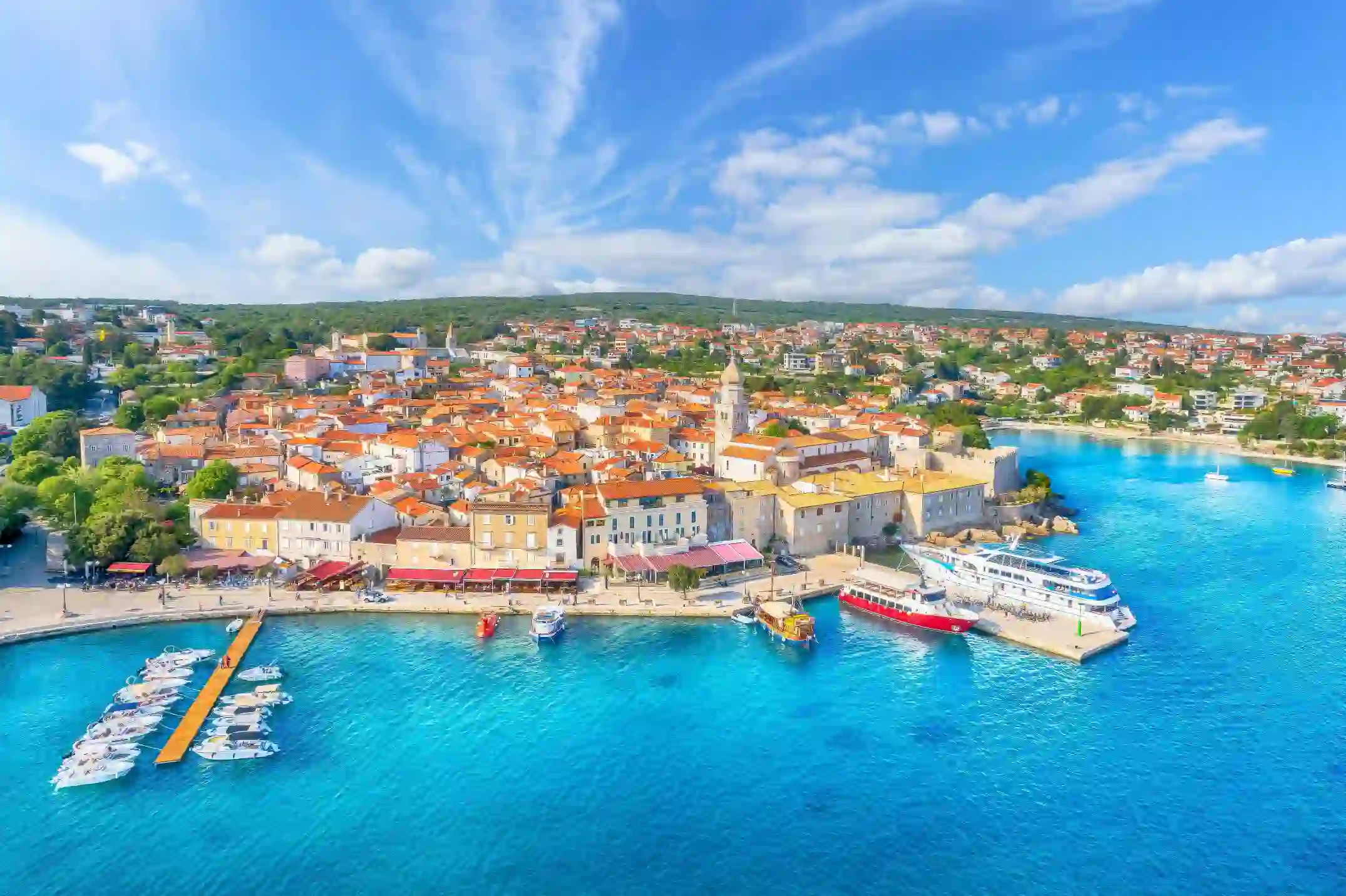 High angle shot of a town on the coast, with bright blue water and boats docked to the left. The town is full of buildings with orange roofs, and the distance is forested land. The sky is blue with some clouds. 