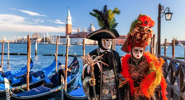 Two people in elaborate Venetian carnival costumes and masks standing by gondolas on a canal, with the Church of San Giorgio Maggiore in the background