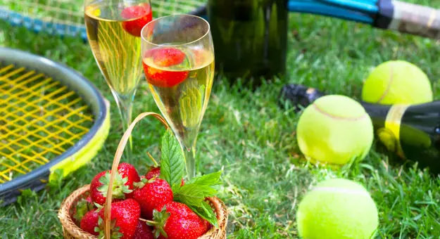 Close up of two glasses of champagne, a basket of strawberries, tennis balls and rackets on some grass