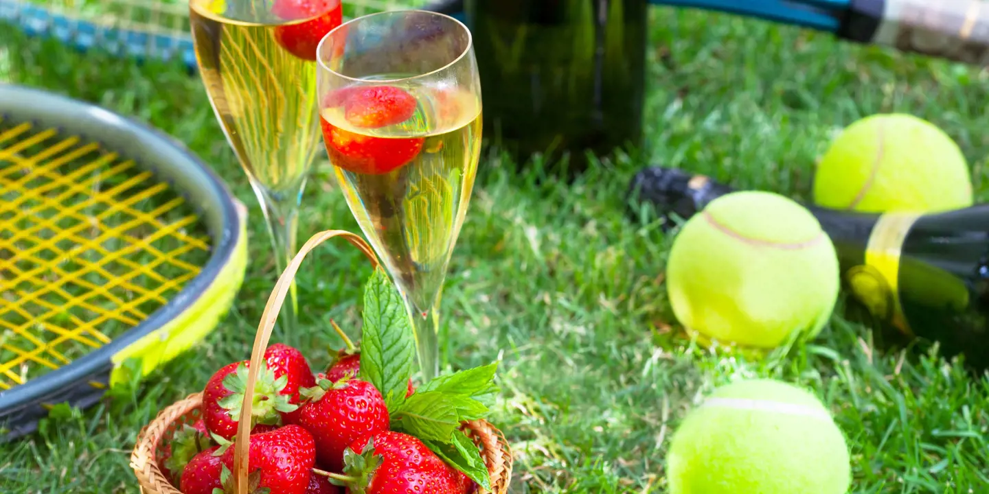 Close up of two glasses of champagne, a basket of strawberries, tennis balls and rackets on some grass