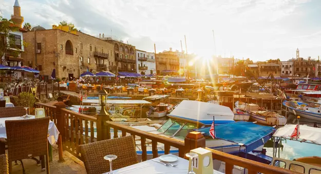 View of a marina from a restaurant, with a laid table in the forefront behind a wooden barrier dividing the boats on the water and the land. Other buildings and restaurants can be seen on the other side, with blue umbrellas and outdoor seating. The sun is poking over the buildings, with beams shining into the camera