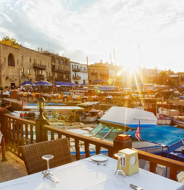 View of a marina from a restaurant, with a laid table in the forefront behind a wooden barrier dividing the boats on the water and the land. Other buildings and restaurants can be seen on the other side, with blue umbrellas and outdoor seating. The sun is poking over the buildings, with beams shining into the camera