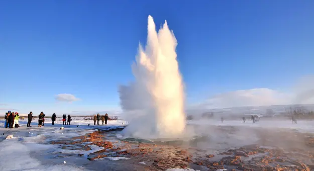 Erupting geysur, surrounded by rocks and ice. People stood watching