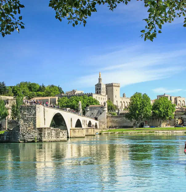 View of the Pont D’Avignon bridge, with the river water in the forefront and a boat coming from the right. In the distance, on the other side of the bridge is a grey palace. The sky is blue.