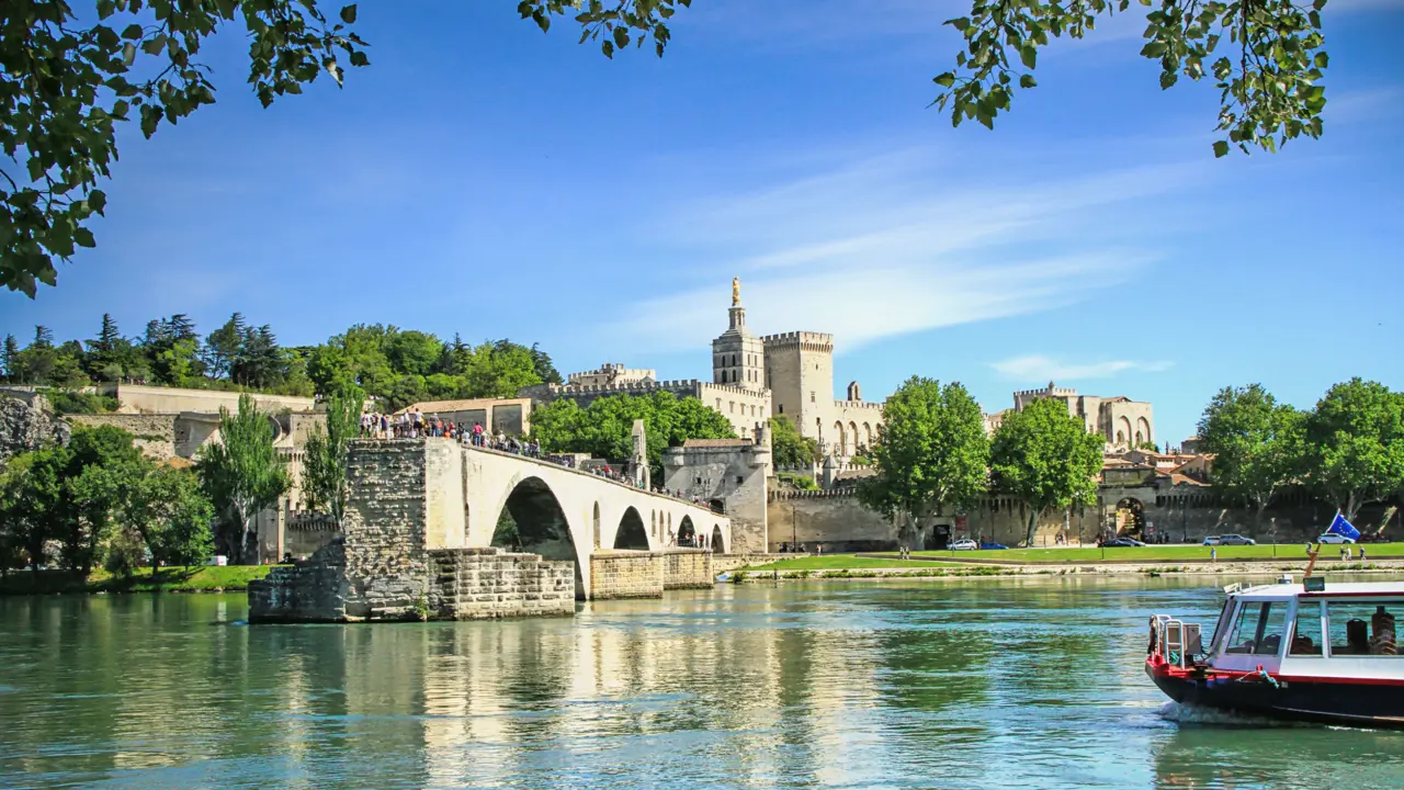 View of the Pont D’Avignon bridge, with the river water in the forefront and a boat coming from the right. In the distance, on the other side of the bridge is a grey palace. The sky is blue.