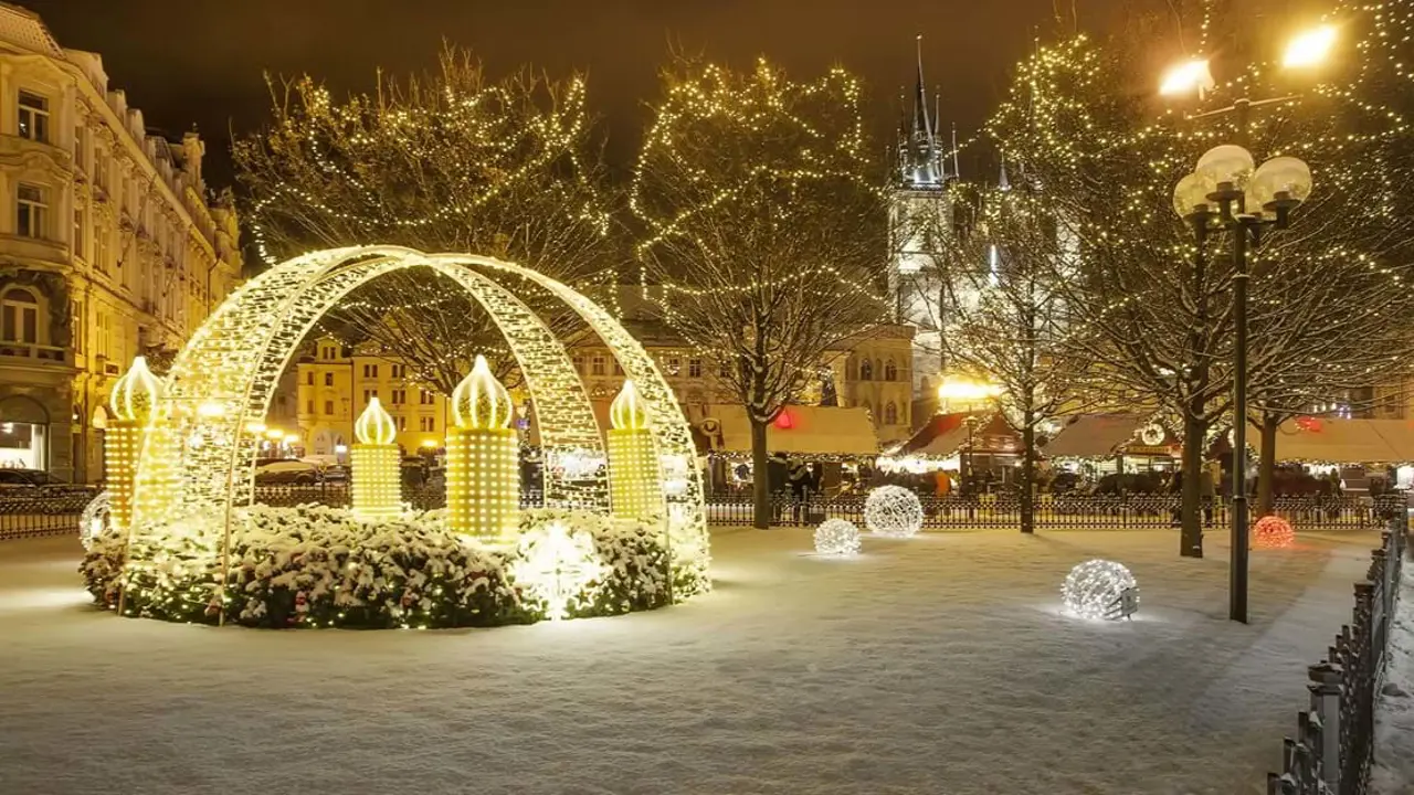 Crossing archways of fairylights with giant lit up artificial candles, fairy lit trees and balls of fairy lights on the snowy grass. A gothic church behind the trees in the distance and a lamp post in the right forefront.