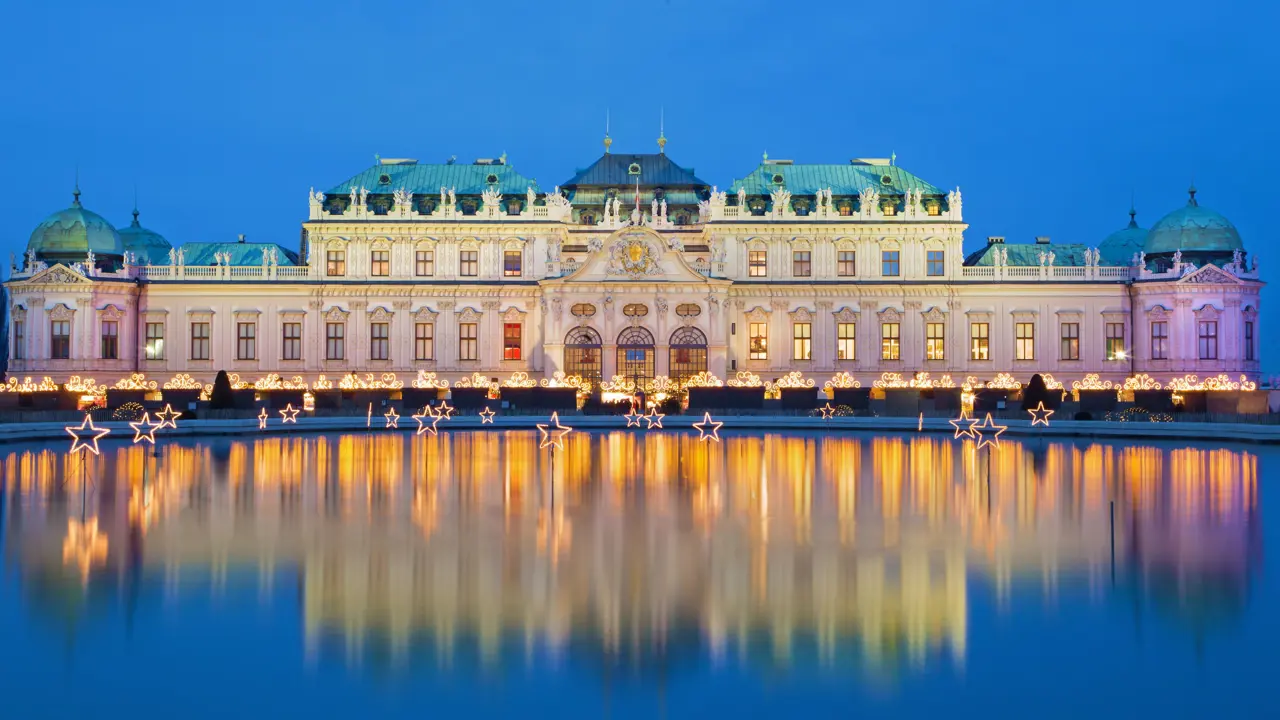 Shot of Schoenbrunn Palace, a wide, beige coloured building with turquoise roofs, in front of a bright blue night sky. The palace has gold, swirly light fixtures along the bottom. In front, is a large pond with lit, stand up stars dotted in.