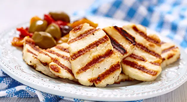 Close up of a dish of grilled halloumi with a side of olives, on a blue and white checkered cloth.