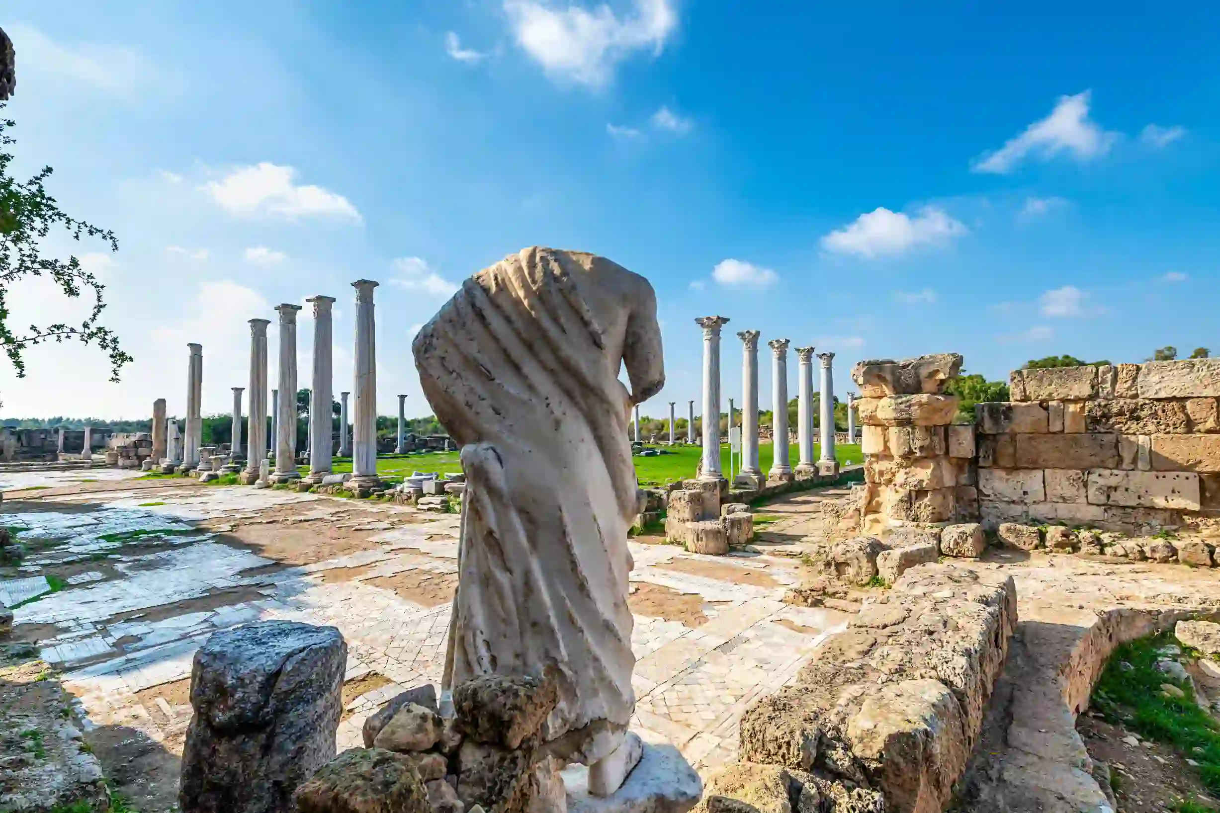 View of Roman ruins, with the back of a headless marble statue of a person. Further away, there is a green lawn surrounded by marble pillars. 