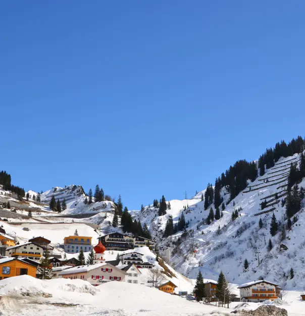 Low-angle view of St Anton village covered in thick snow, with a few houses and hotels going up the mountain and fir trees dotted all over. In front of a blue sky.