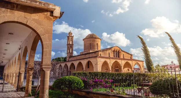 View of a beige stone building, archways round the bottom, a triangular roof, dome shaped tower, and a thin tower. In front is a gated garden with bushes, trees and flowers. To the left is a beige stone archway