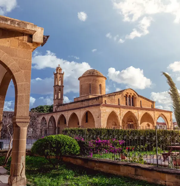 View of a beige stone building, archways round the bottom, a triangular roof, dome shaped tower, and a thin tower. In front is a gated garden with bushes, trees and flowers. To the left is a beige stone archway