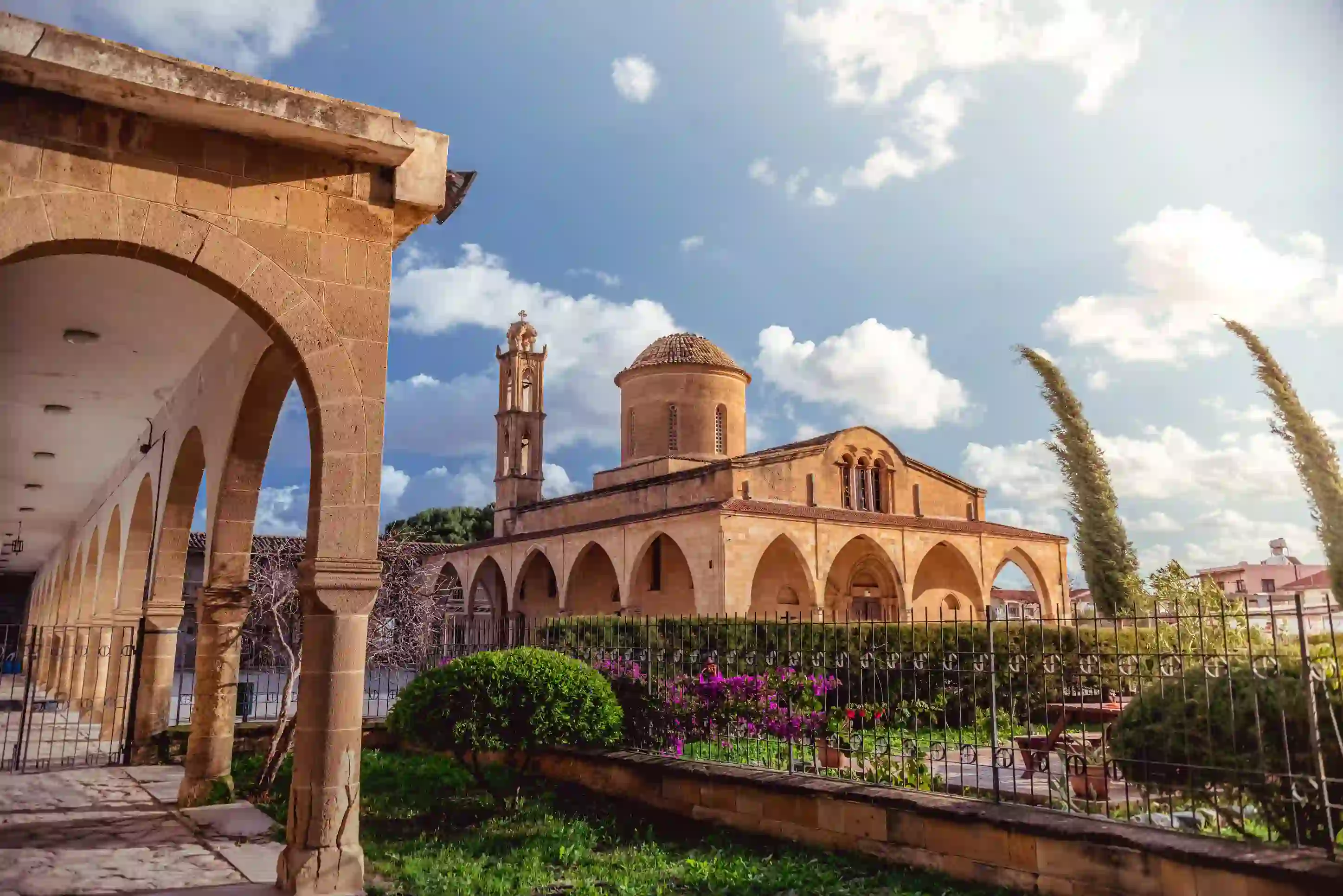 View of a beige stone building, archways round the bottom, a triangular roof, dome shaped tower, and a thin tower. In front is a gated garden with bushes, trees and flowers. To the left is a beige stone archway