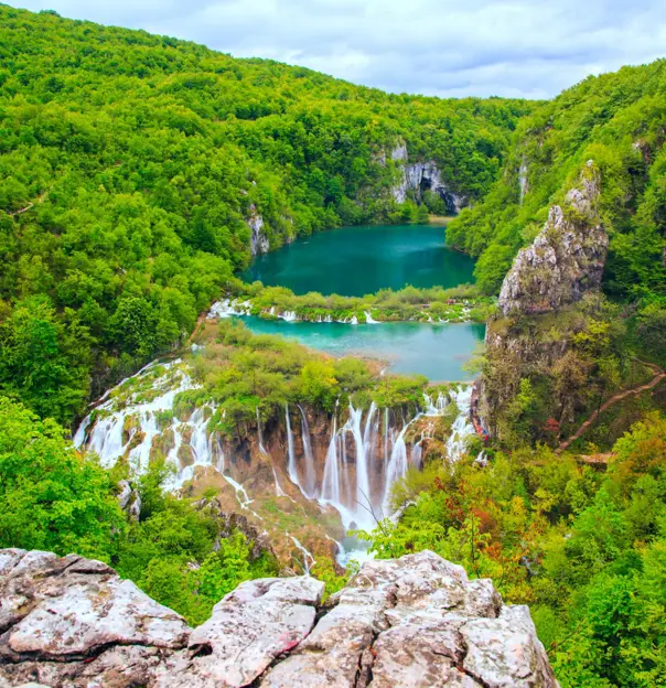 High shot of waterfalls in front of a dark turquoise body of water, surrounded by forested hills. In the forefront is a piece of rock and trees.