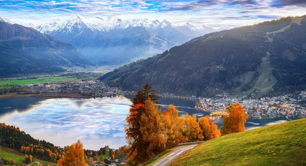 View of Austrian mountains, with some in the distance with snow on the top. Below is a town where small buildings can be seen, behind a lake. To the right another town, and in the forefront, orange autumnal trees and a grassy hill and a pathway