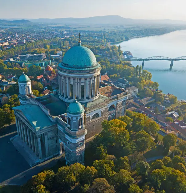 Bird's eye view of the Basilica of Esztergom, with it's turquoise domed roof with a cross on in the centre, and two smaller towers at the front with domed turquoise tops. Danube river in the background, with a bridge going across.