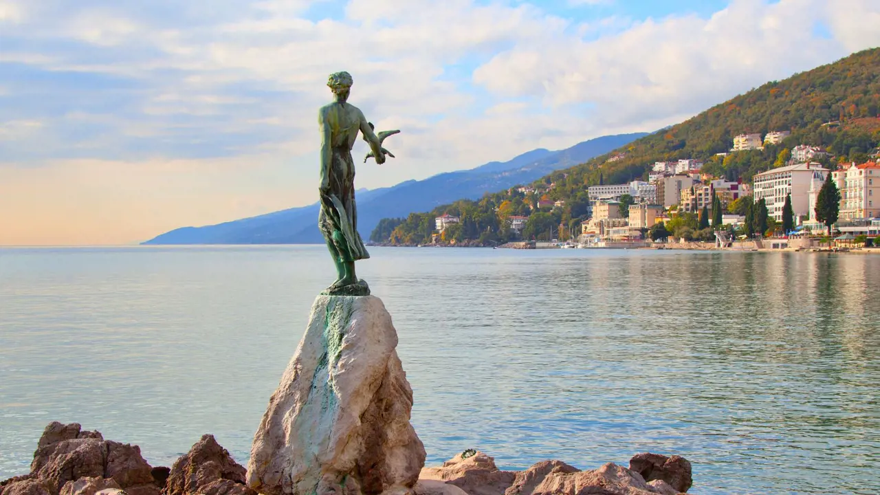 Back of a green statue of a lady holding a bird, on some rocks, facing the sea. Past the sea, there is a silhouette of a mountain, and on the right is a town. The sky is blue but cloudy